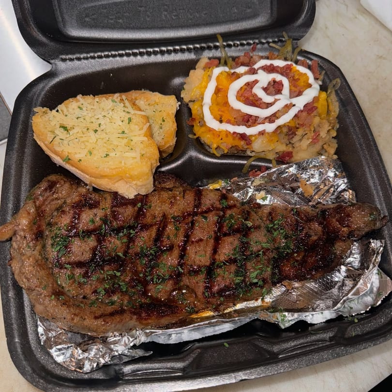 Smiley's Grill steak dinner with garlic bread and loaded mashed potatoes in a takeout container.