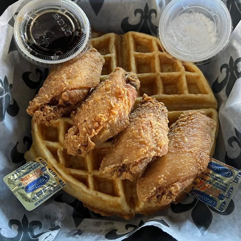 Crispy fried chicken wings and a waffle, with syrup, butter, and powdered sugar, in a basket.