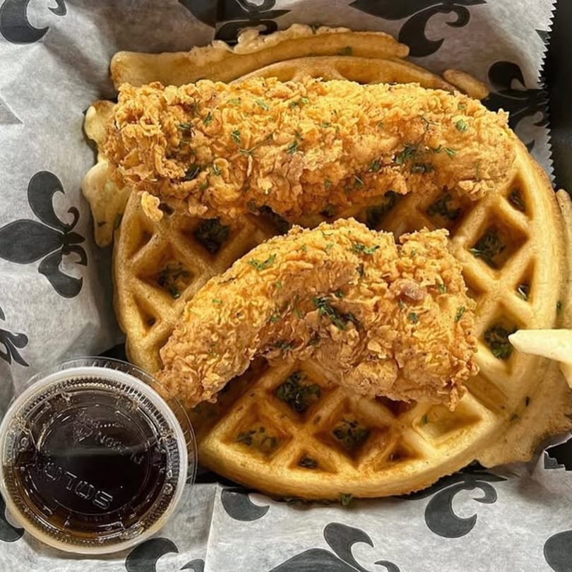 Crispy fried chicken tenders on a waffle with a side of syrup, served on fleur-de-lis patterned paper.