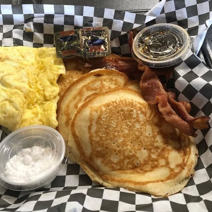 Breakfast platter with scrambled eggs, pancakes, bacon, and syrup in a checkered paper-lined basket.